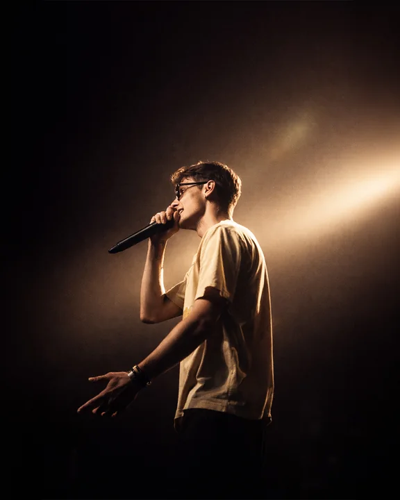 Un cantante tiene un microfono sotto un fascio diagonale di luce calda su un palco buio.