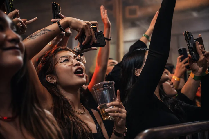 Il pubblico di un concerto alza mani e telefoni, con una persona con gli occhiali che canta in primo piano.