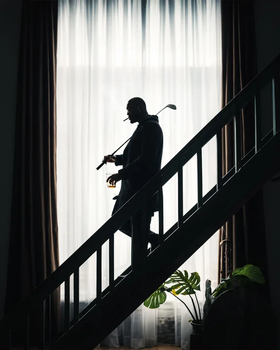 Una persona in silhouette porta una mazza da golf e un bicchiere su una scala accanto a un'alta finestra con tende.