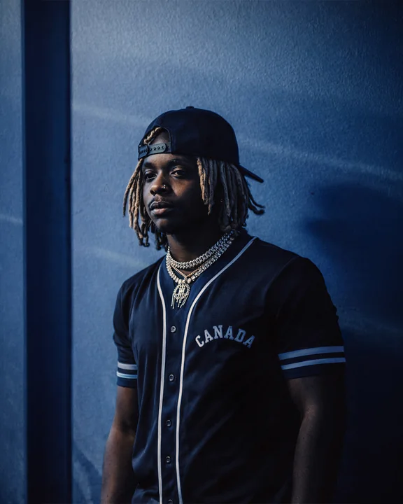 Un uomo con dreadlock, berretto girato all'indietro e collane sovrapposte è in piedi contro una parete blu, con una maglia da baseball scura.