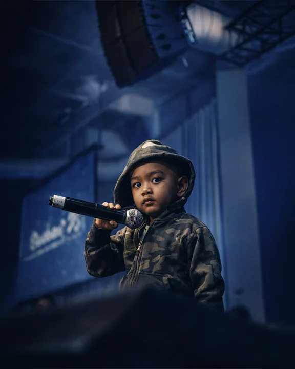 Un bambino con cappello e giacca mimetici tiene un microfono sotto luci da palco blu.