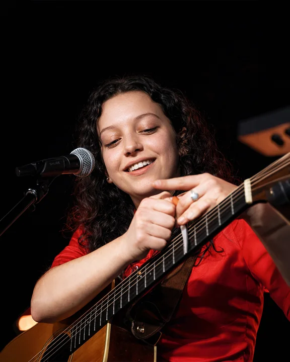 Un musicista sorridente con una maglia rossa tiene una chitarra acustica accanto a un microfono, sullo sfondo scuro di un palco.