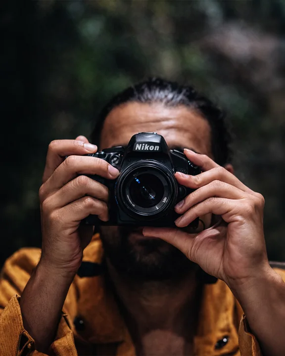 Una persona con una maglia color senape tiene una macchina fotografica direttamente davanti al viso, all'aperto.