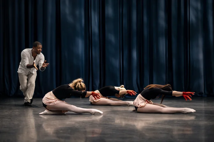 Tre ballerine con gonne rosa pallido e guanti rossi si protendono in avanti dal pavimento del palco, mentre un altro artista gesticola alle loro spalle contro tende blu.