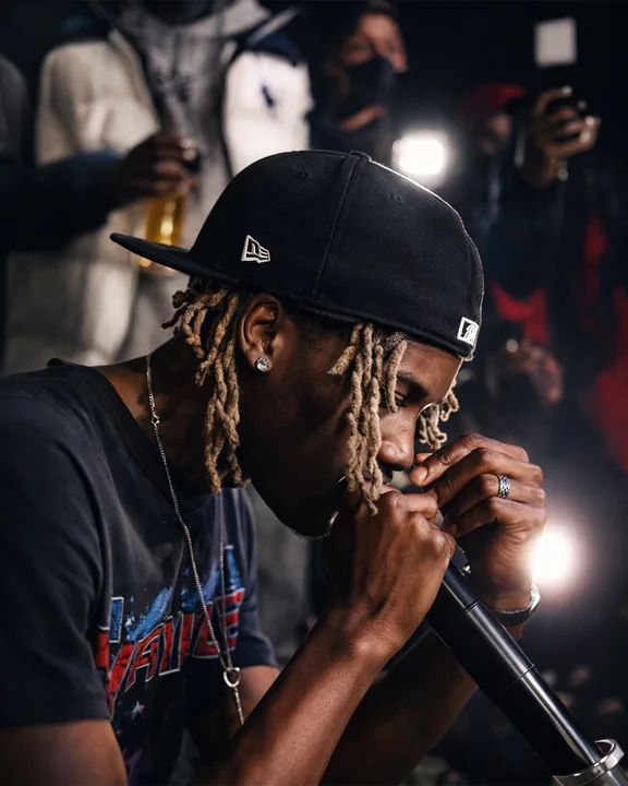Un artista con dreadlock chiari e un berretto nero girato all'indietro si avvicina a un microfono, mentre alle sue spalle alcune persone alzano i telefoni.