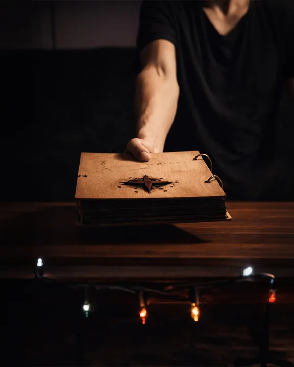 Eine Person reicht ein ringgebundenes Buch mit Holzdeckel über einen Tresen, der von kleinen bunten Lichtern beleuchtet wird.
