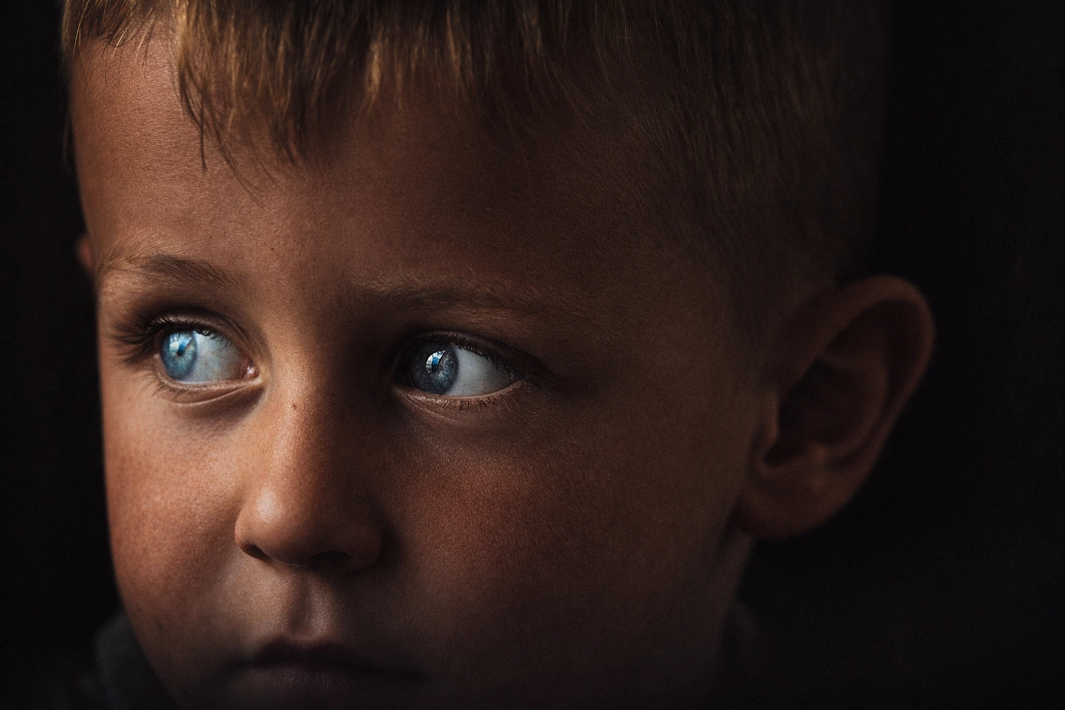 Close portrait of a child with blue eyes looking sideways, with warm light falling across the face.