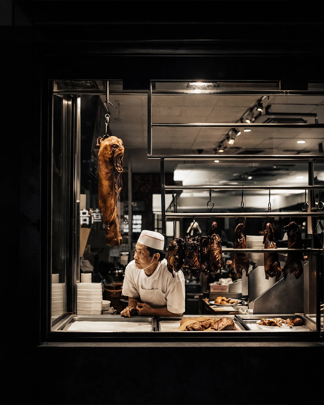 A cook in a white hat and uniform works behind the window of a brightly lit food counter.