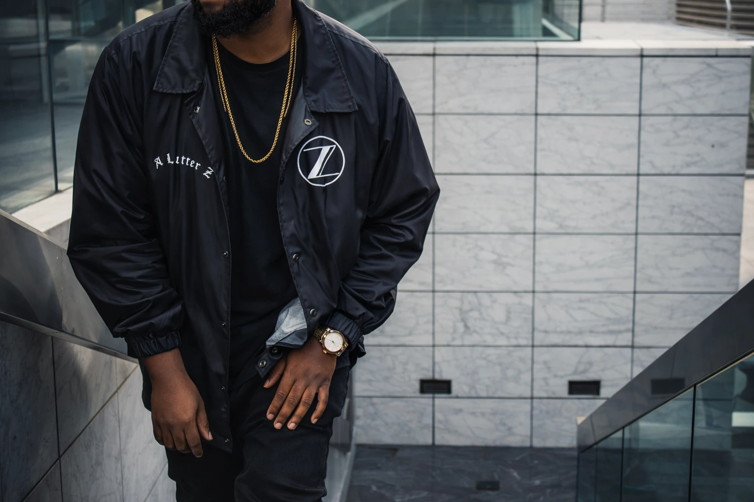 A cropped street portrait showing a dark jacket, gold chain and watch against grey tiled architecture.
