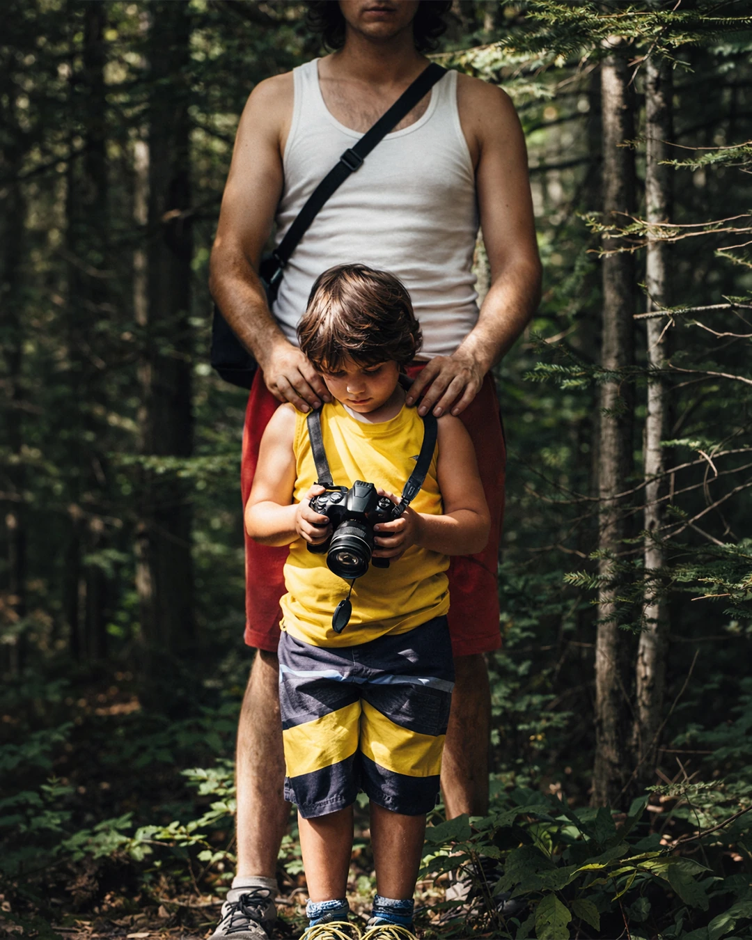 A child holds a camera on a wooded trail while an adult stands behind with hands on the child’s shoulders.