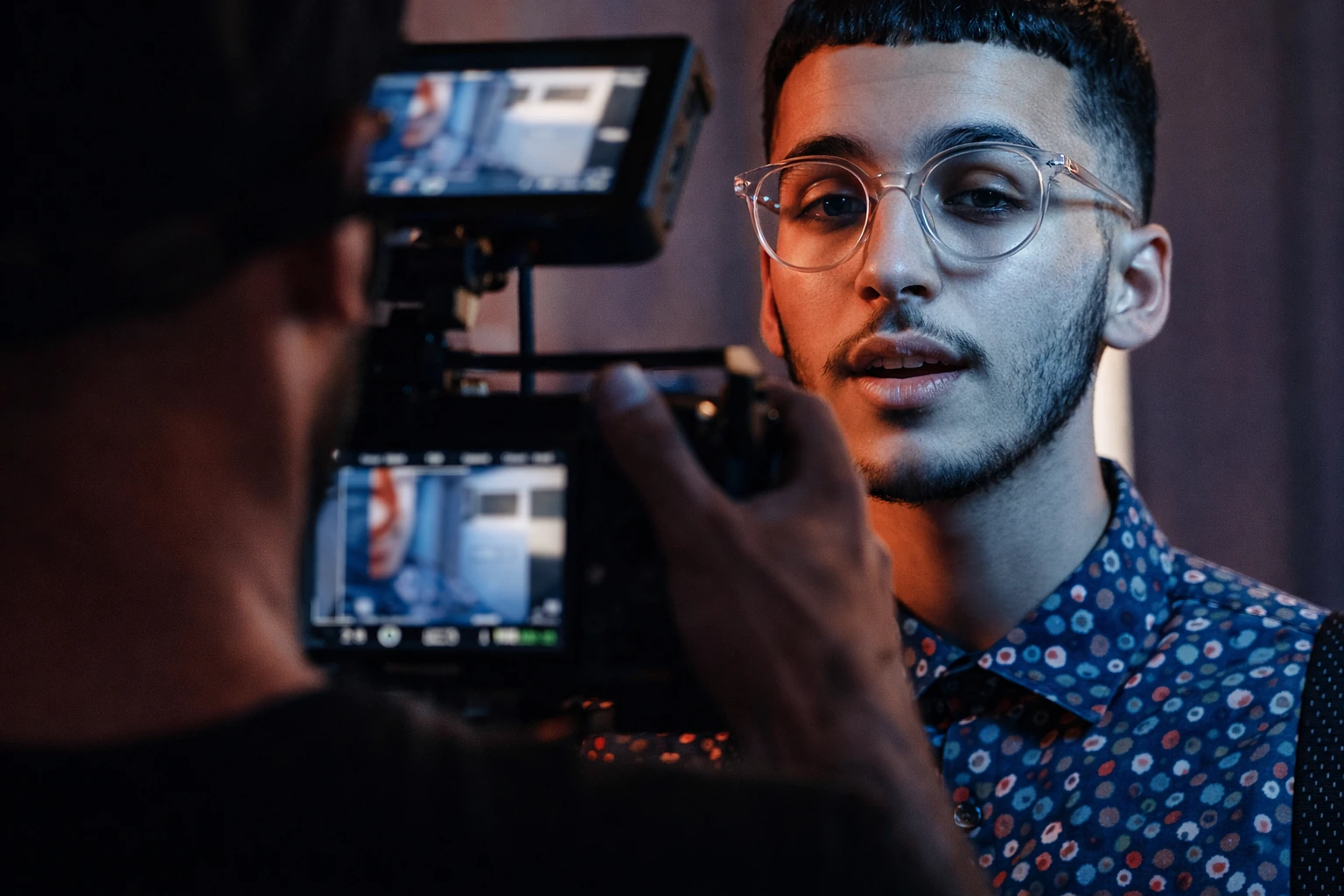 A camera in the foreground frames a man with glasses and a patterned shirt in blue and red light.