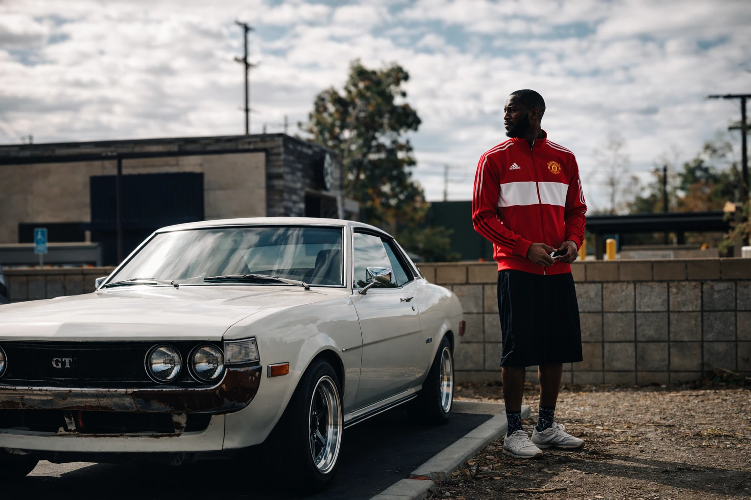 A man in a red track jacket stands beside a white vintage car beneath a cloudy sky.