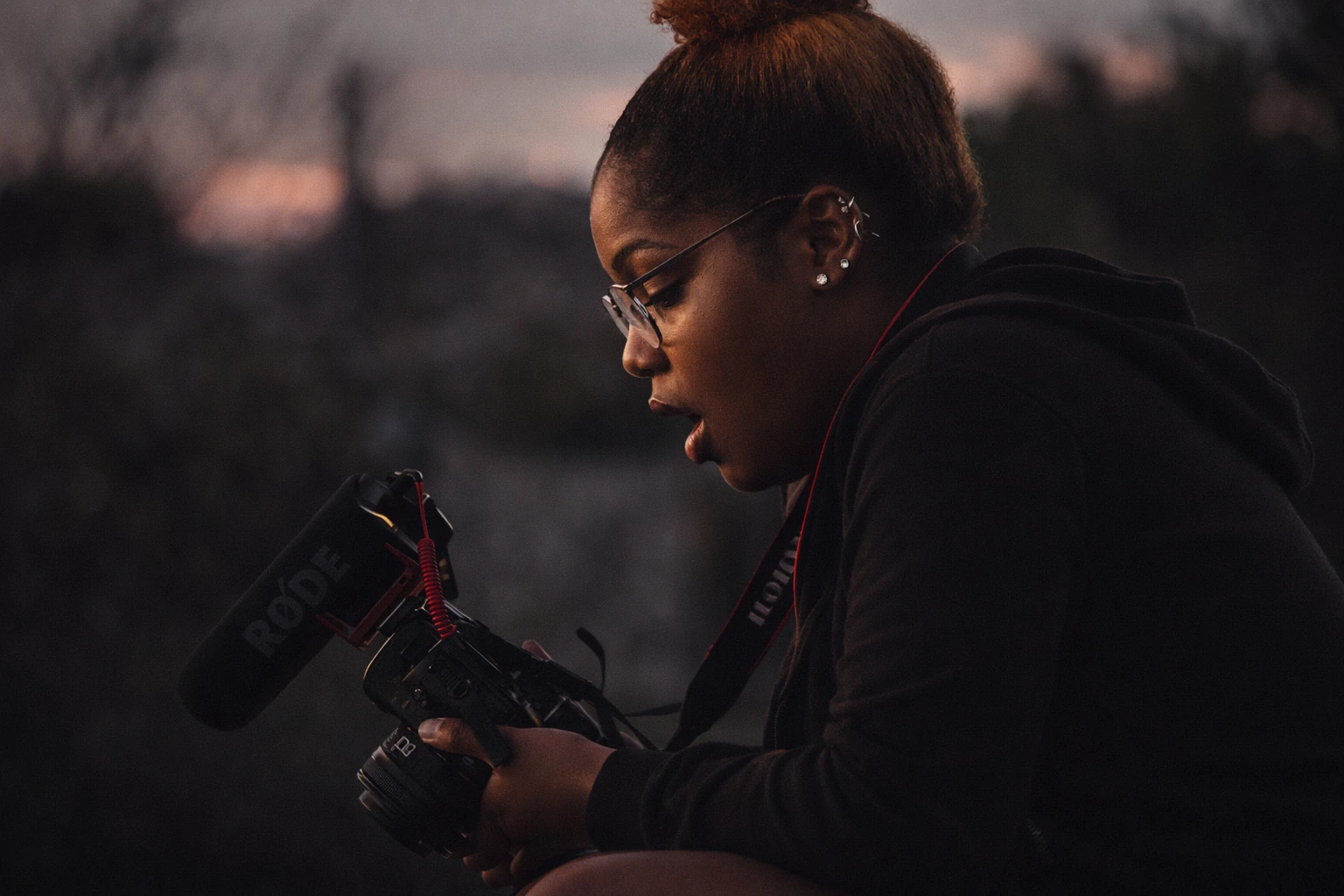 A camera operator with glasses and hair in a bun works with a cinema camera outdoors at dusk.