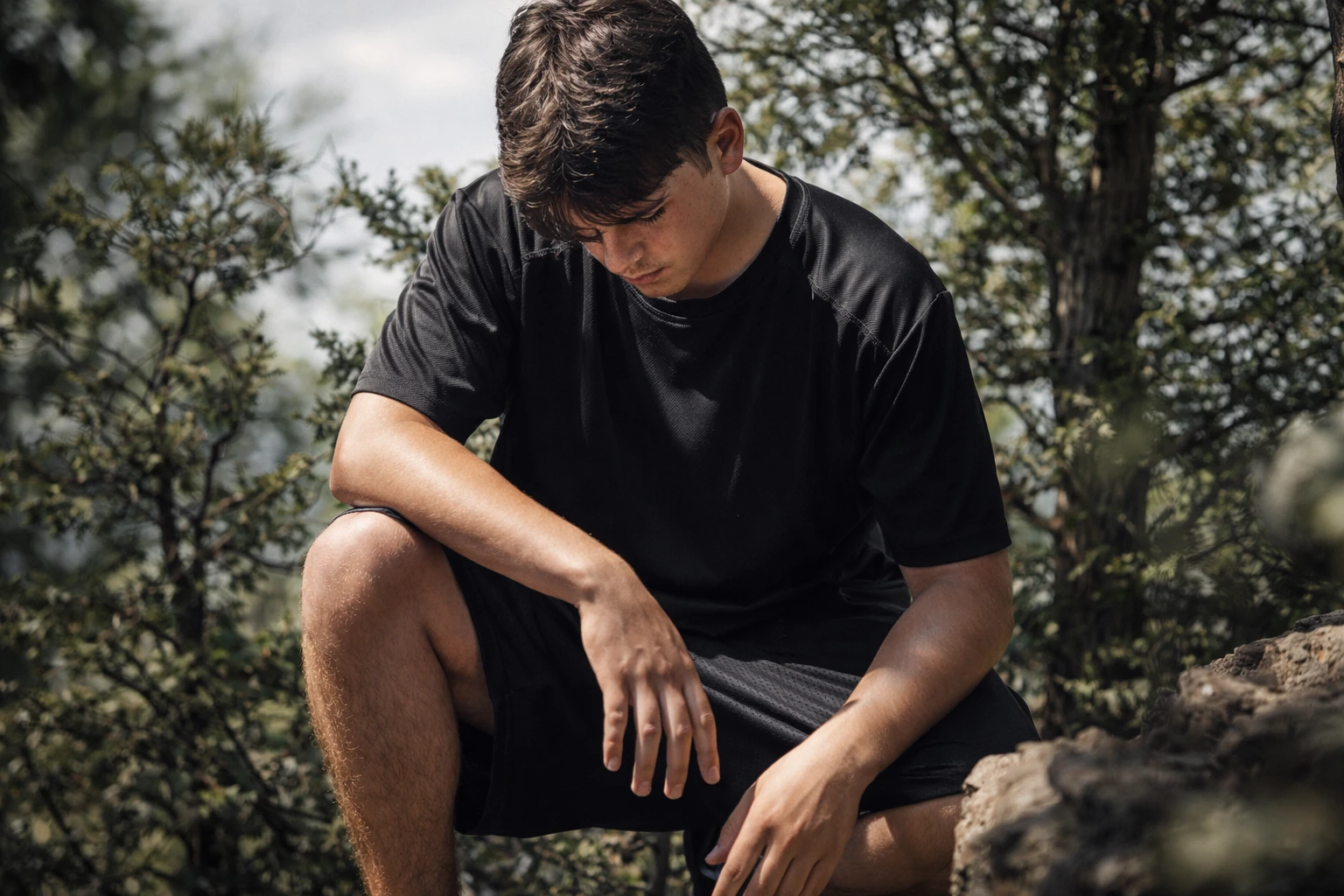 A person in a black T-shirt and shorts sits among rocks and trees with their head lowered.