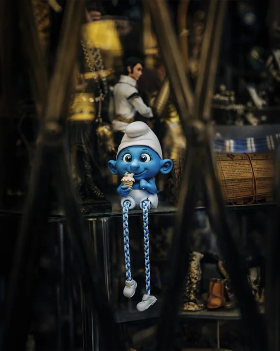 Eine blaue Schlumpffigur mit weißer Mütze und langen Beinen aus Schnur hängt zwischen alten Gegenständen in einem Laden.