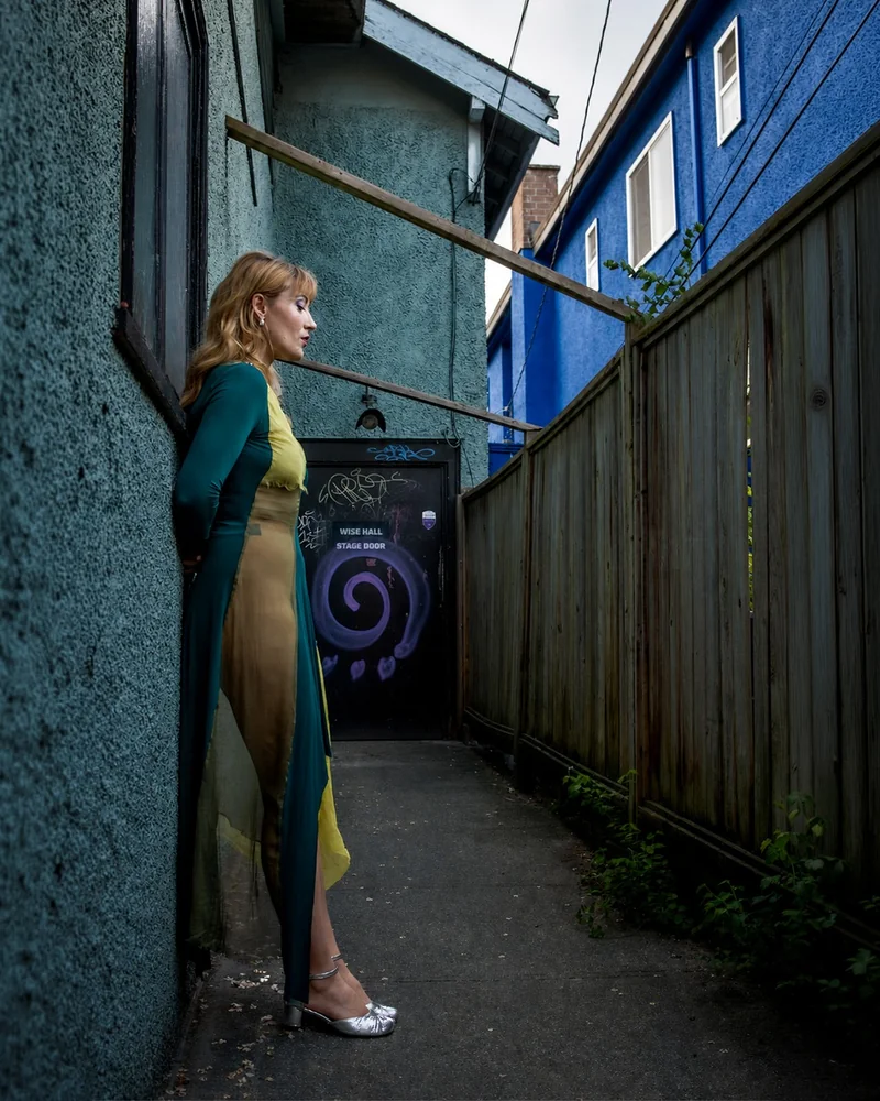 Una donna con un lungo abito verde e oro è in piedi in uno stretto passaggio fra una parete materica, una recinzione di legno e un edificio blu acceso.