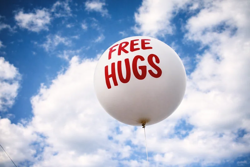Un grande palloncino bianco con la scritta FREE HUGS in lettere rosse galleggia contro un cielo azzurro e morbide nuvole bianche.