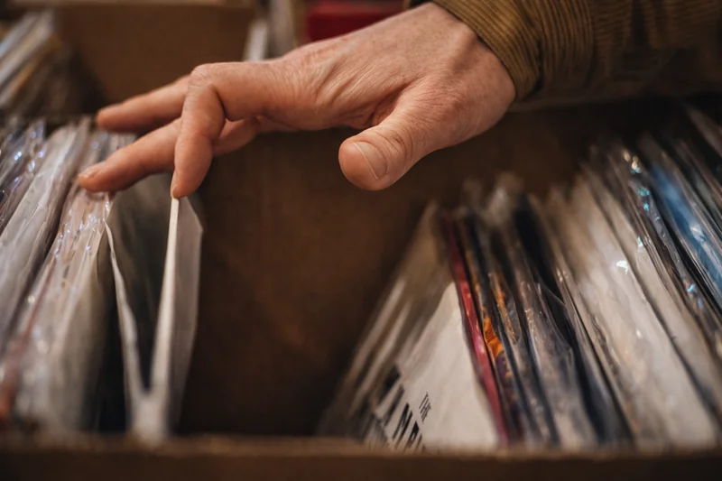 Una mano si allunga fra dischi in vinile nelle loro buste protettive dentro un espositore di legno.