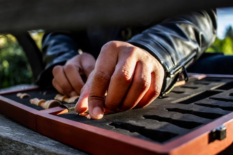 Sonnenbeschienene Hände greifen unter den Ärmeln einer schwarzen Lederjacke in einen passgenauen Holzkasten mit hellen Schachfiguren.