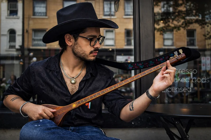 Un musicista con un cappello nero a tesa larga e occhiali suona uno strumento a corde di legno dal lungo manico accanto alla vetrina di un caffè.