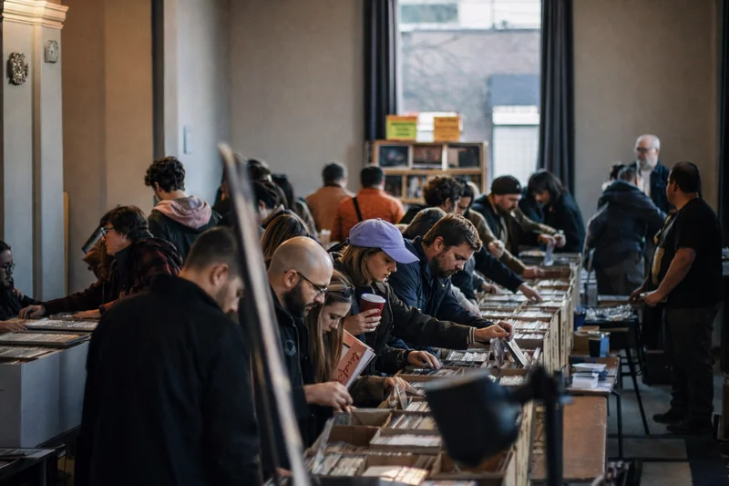 Persone sfogliano lunghe file di espositori di dischi in vinile in una sala luminosa con alte finestre.