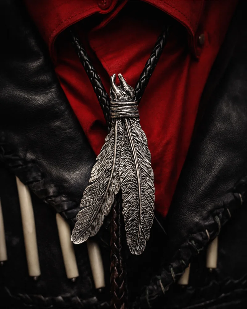 Un ciondolo d'argento a forma di due piume pende sopra una maglia rossa e un gilet di pelle nera.