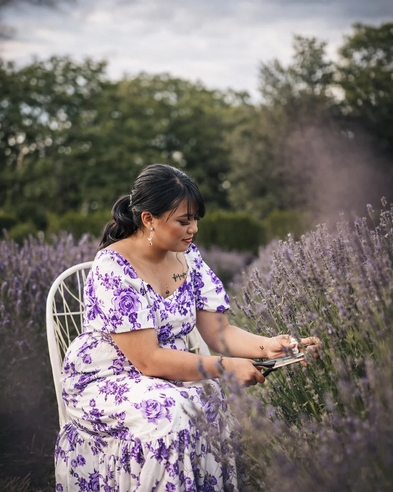 Una donna con un abito floreale bianco e viola siede su una sedia bianca e taglia steli di lavanda con le forbici.