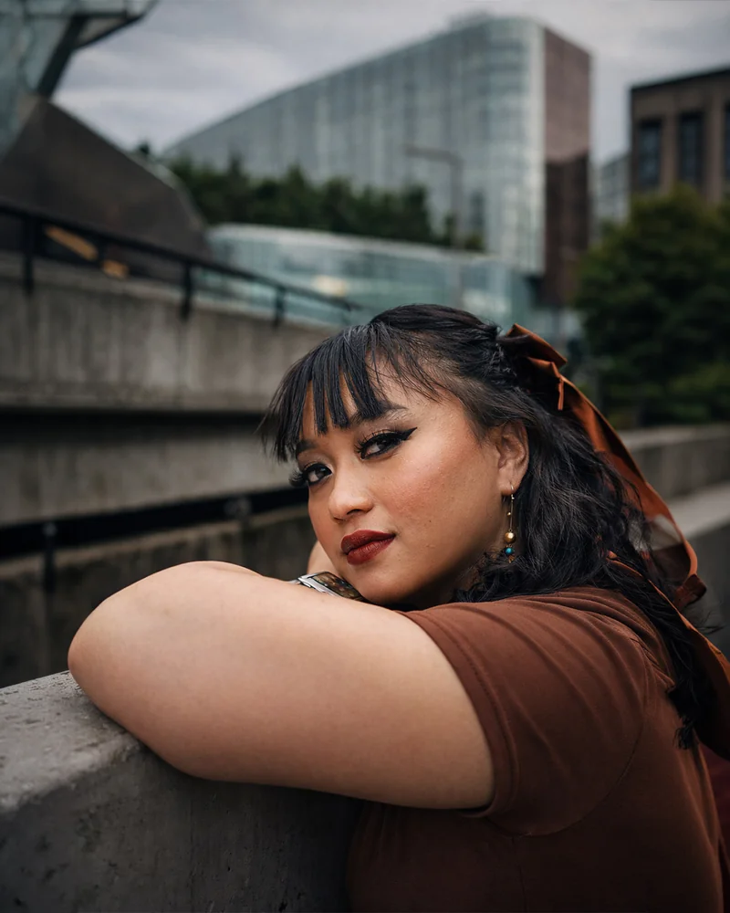 Una donna con capelli scuri e frangia appoggia le braccia su una balaustra di cemento e guarda verso la macchina fotografica, con edifici cittadini alle spalle.