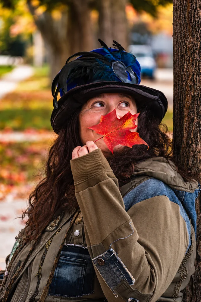 Eine Person mit dunklem Federhut hält sich neben einem Baum ein rotes Ahornblatt vor den Mund und schaut nach oben.