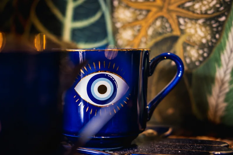 Eine kobaltblaue Tasse mit gemaltem Auge und goldenen Strahlen steht auf einer dunklen Untertasse; dahinter sind Gefäße weich verschwommen.