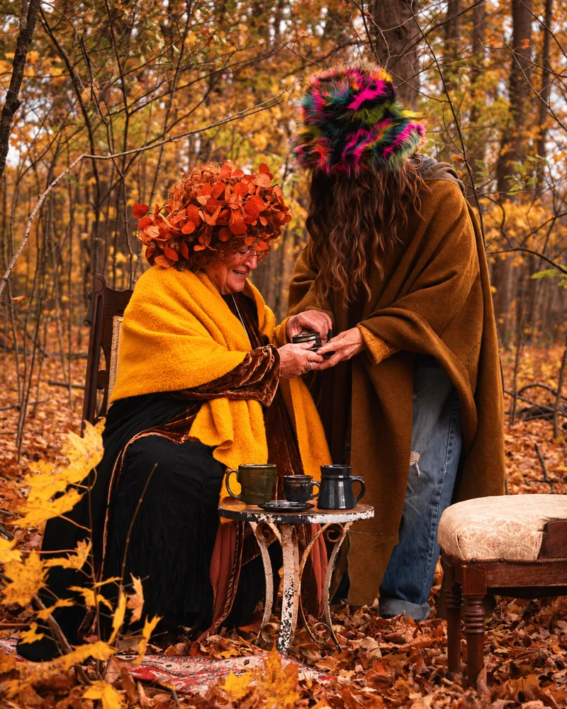 Zwei Menschen mit aufwendigen Hüten und warmen Umhängen teilen im herbstlichen Wald neben einem kleinen Tisch voller Becher eine Tasse.