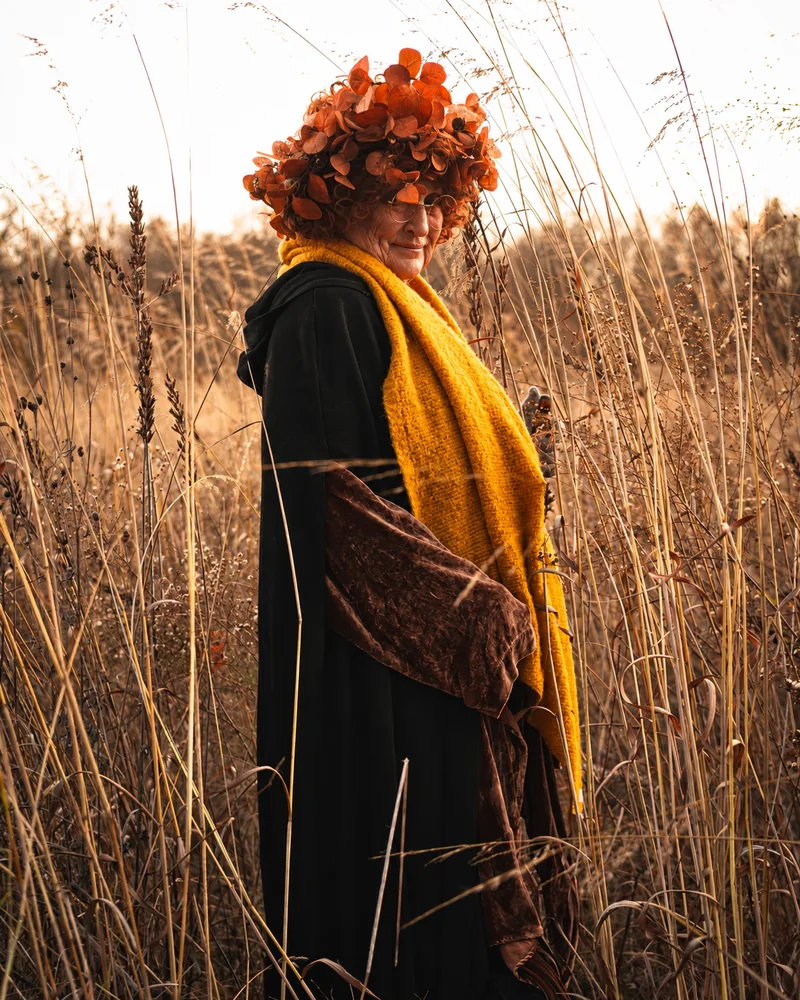 Eine Person mit Brille, aufwendigem orangefarbenem Blumenhut, gelbem Schal und dunklem Umhang wendet sich zwischen hohen goldenen Schilfhalmen der Kamera zu.