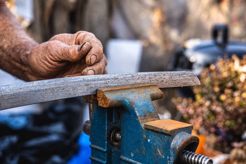 Eine von Arbeit gezeichnete Hand hält ein kleines Werkzeug über einem Metallstreifen, der in einem verwitterten blauen Schraubstock eingespannt ist.