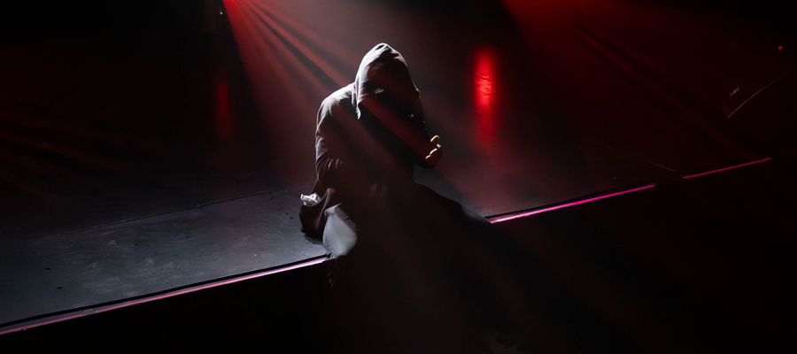 Solitude in the Spotlight A lone, hooded figure sitting on the edge of a dark stage illuminated by a single, dramatic red spotlight.