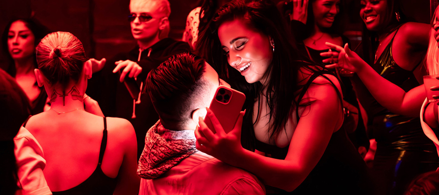 Reflections of the Night A smiling woman holding up a smartphone to take a photo of a man in a red-lit, crowded club setting.