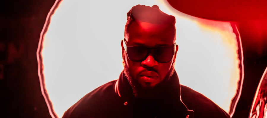 The Eclipse A portrait of a man wearing sunglasses, intensely backlit by a bright circular light against a deep red backdrop.