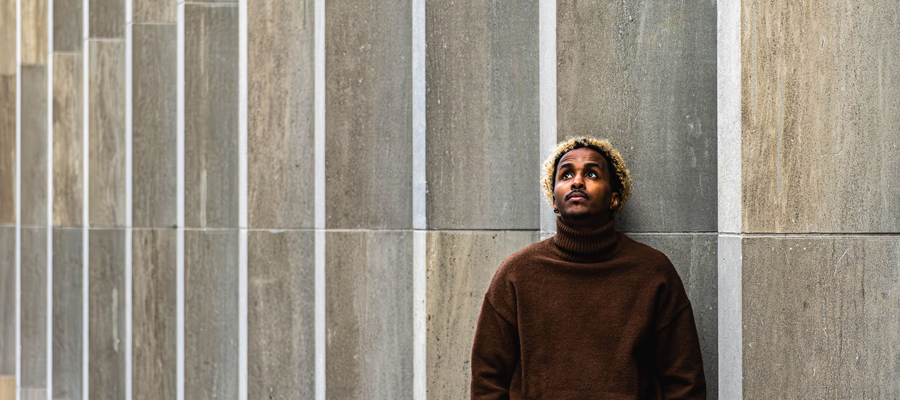 Vertical Horizons A man in a brown turtleneck leaning against a modern, vertically paneled concrete wall, looking thoughtfully upward.