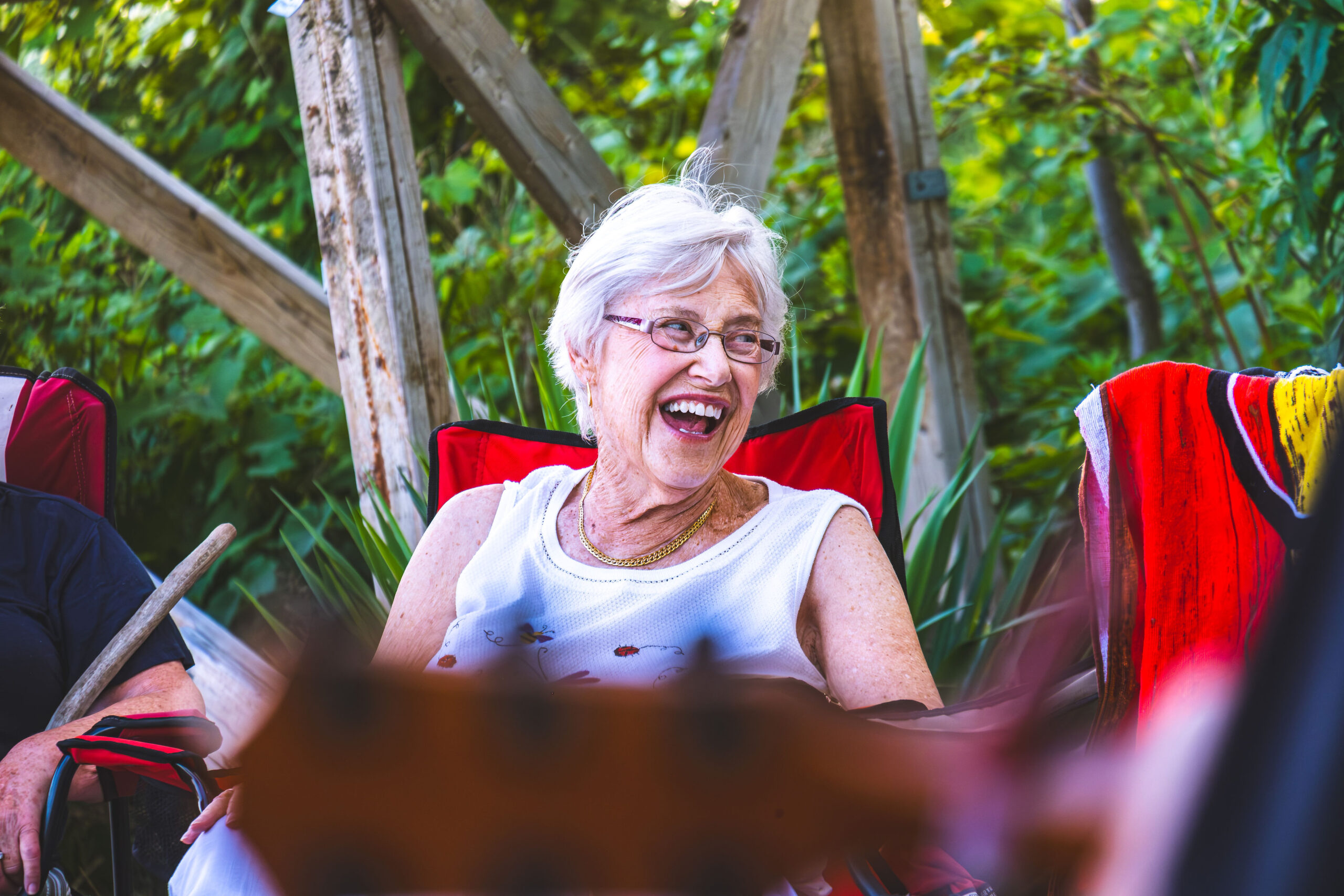 An elderly woman with white hair laughing joyfully while sitting in a red camping chair outdoors.