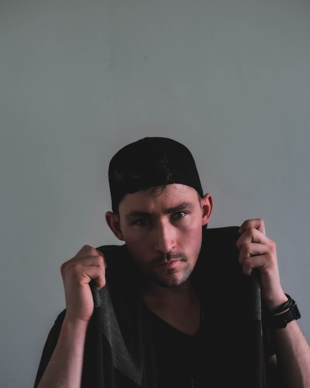 Moody portrait of a young man wearing a backwards black cap, pulling his dark collar up against a grey background.