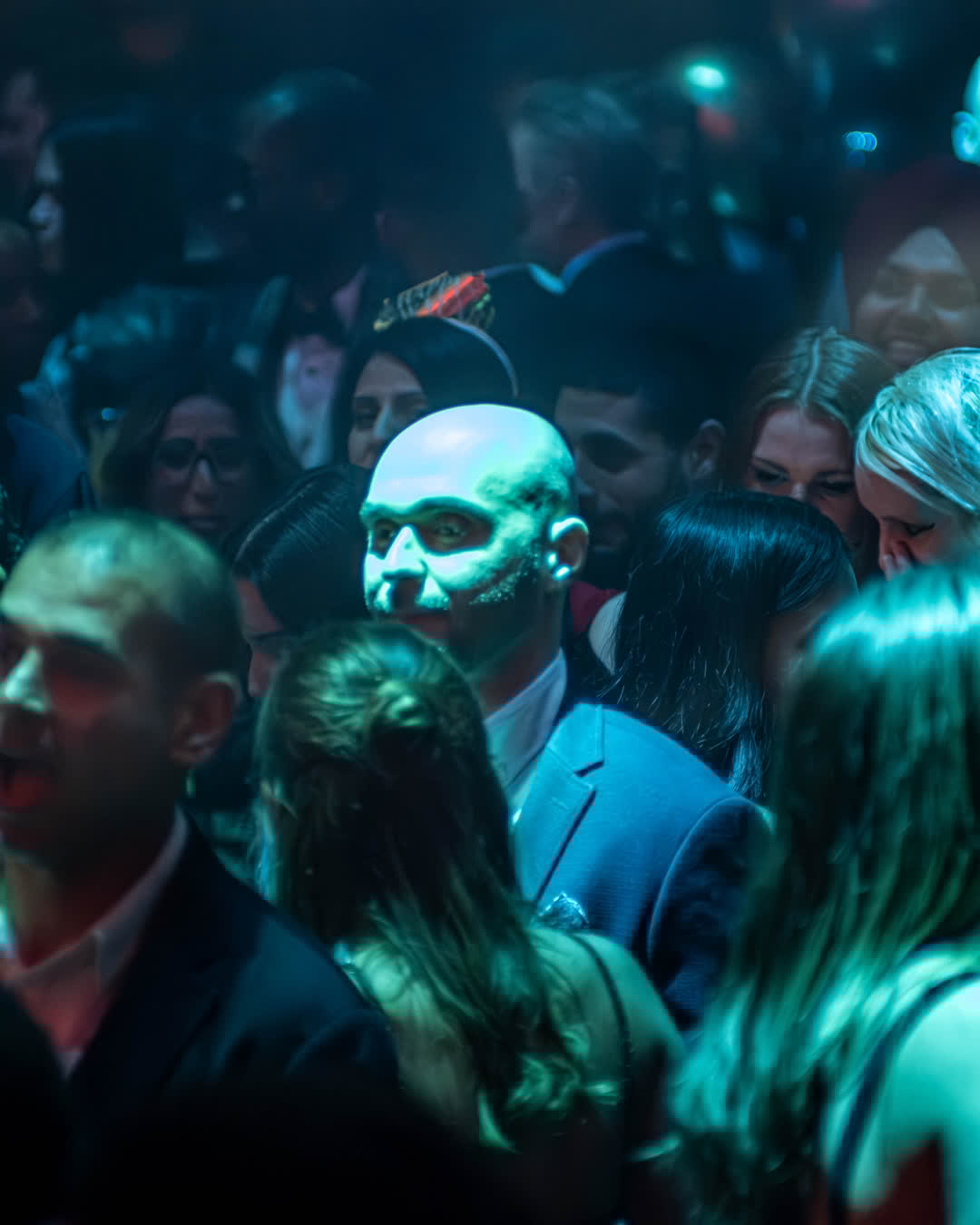 A crowded nightclub scene with a bald man's face illuminated by bright teal and blue lights amidst the dark crowd.