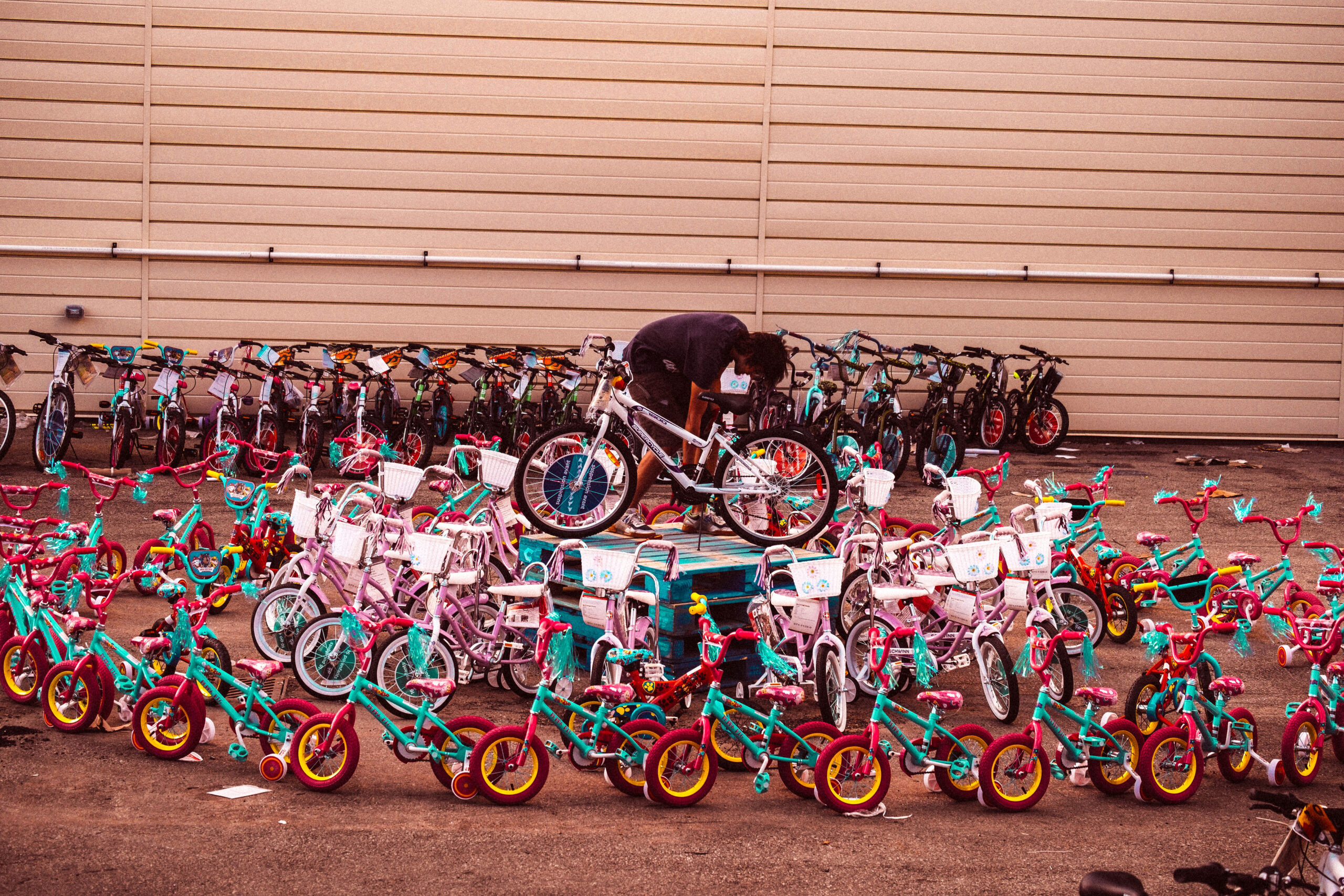 Dozens of brightly colored children's bicycles surrounding a wooden pallet where a person is assembling a white bike outdoors.