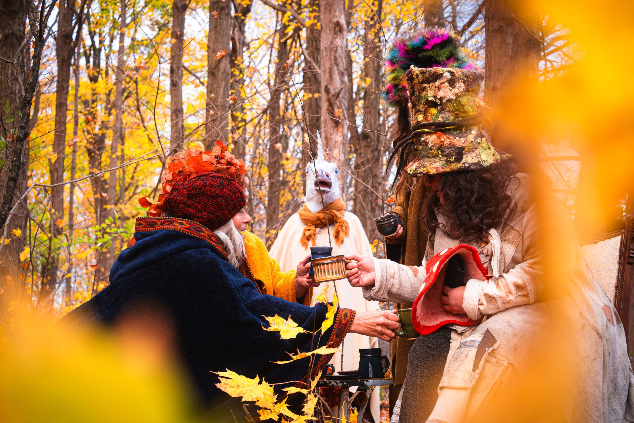 A whimsical autumn gathering in the woods featuring people in elaborate fantasy costumes and a person wearing a white unicorn mask drinking from mugs.