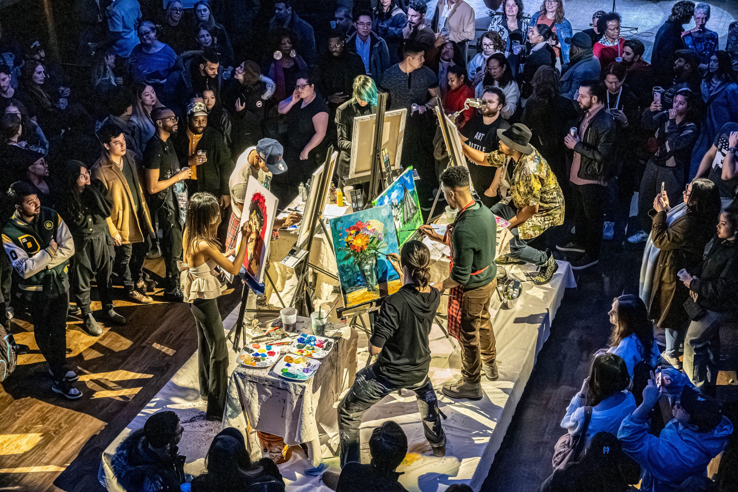 High-angle view of an indoor live art competition where artists paint on easels surrounded by a large crowd of spectators.