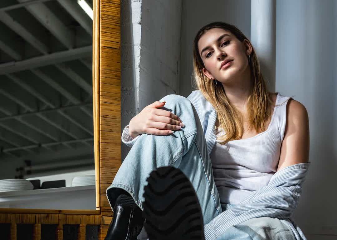A young woman sitting on the floor in a bright studio, leaning against a white pillar, wearing jeans and black boots, with a large bamboo mirror behind her.