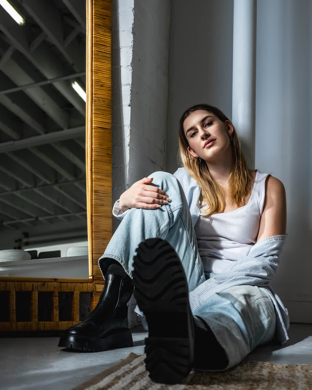A young woman sitting on the floor in a bright studio, leaning against a white pillar, wearing jeans and black boots, with a large bamboo mirror behind her.