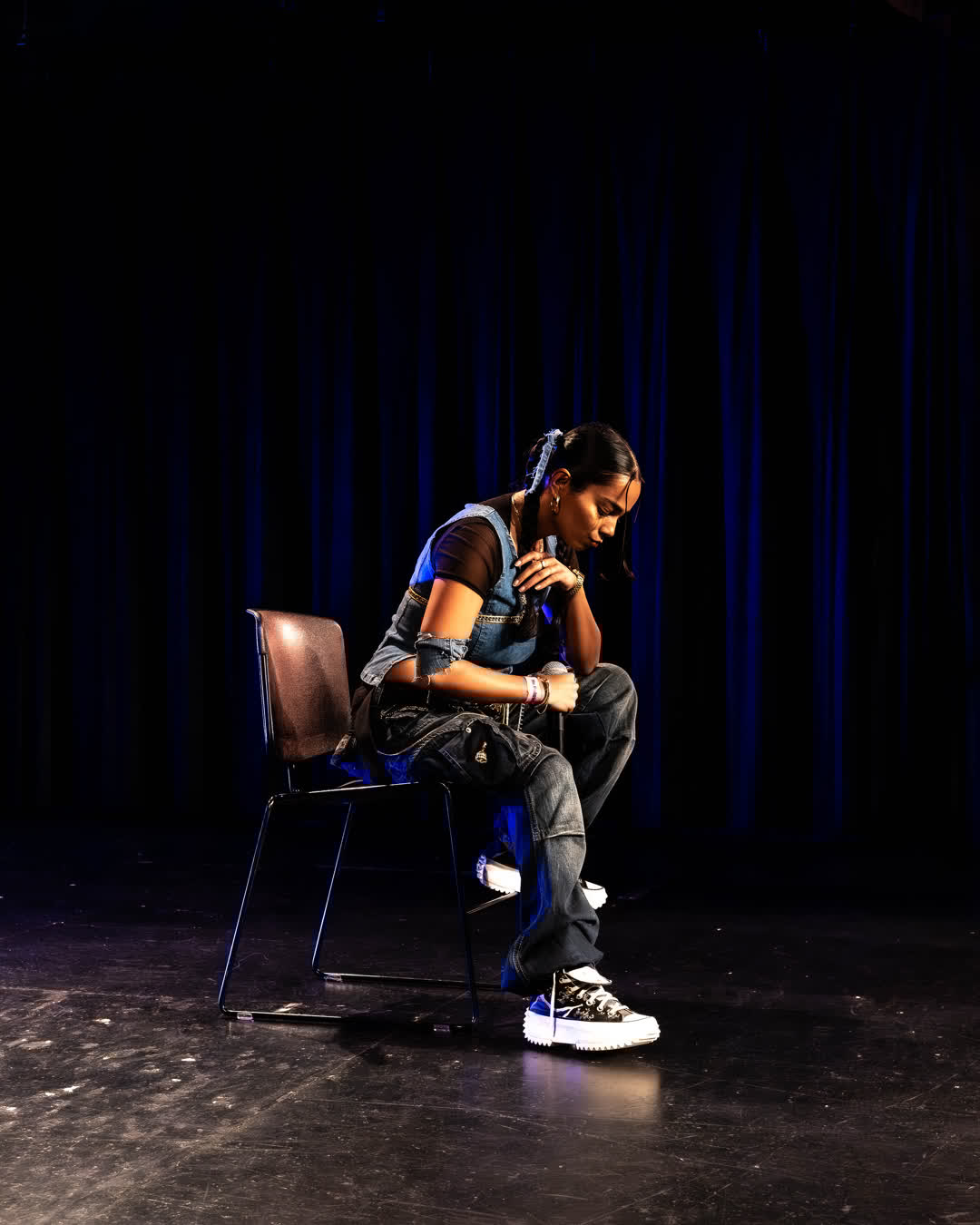 A young woman in a denim outfit sitting thoughtfully on a chair on a dark stage holding a microphone with blue curtains in the background.