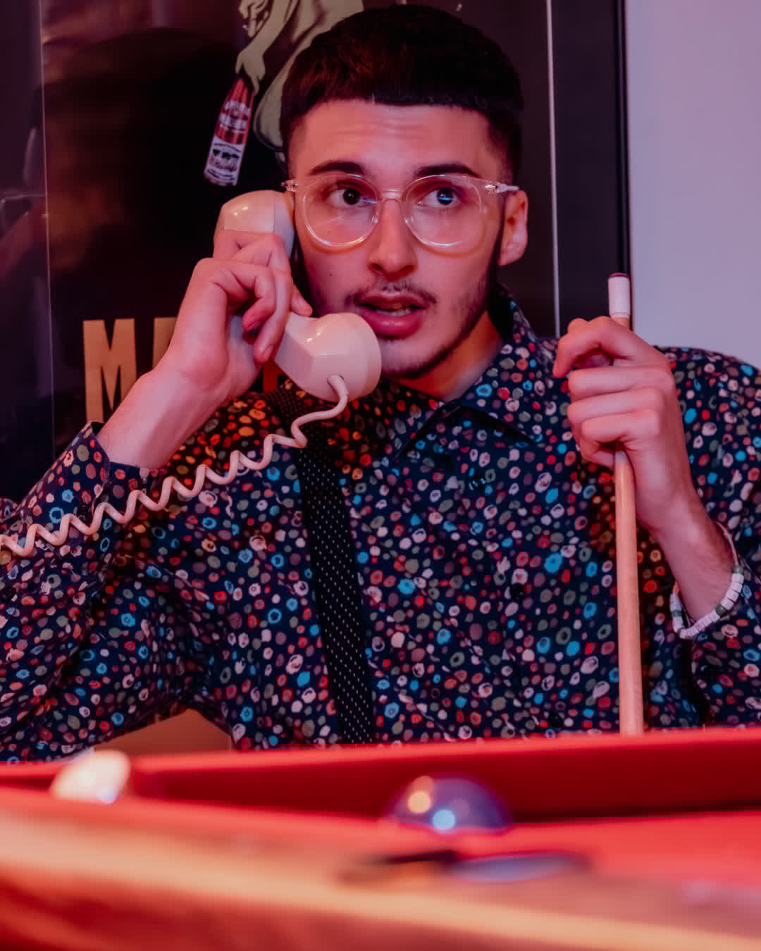 A young man with clear glasses and a patterned shirt speaking on a retro white telephone while holding a pool cue in a dim, moody room.