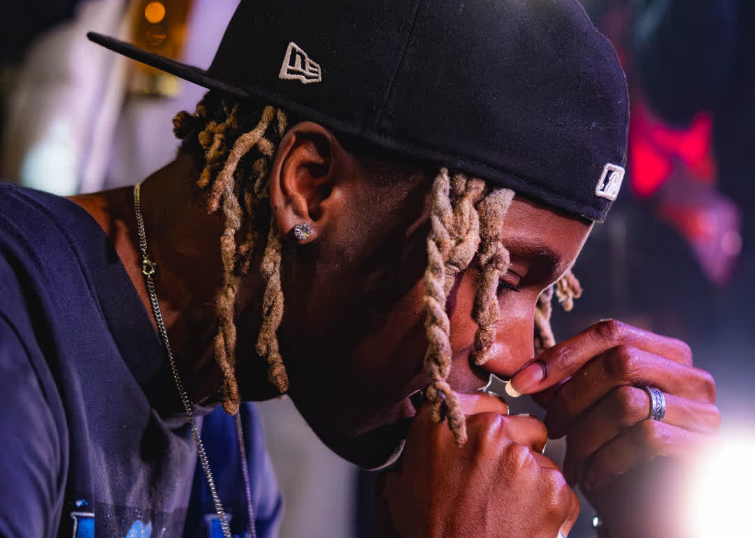 A musical artist with dreadlocks and a black fitted cap holding a microphone close to his face during a live, intensely lit performance.