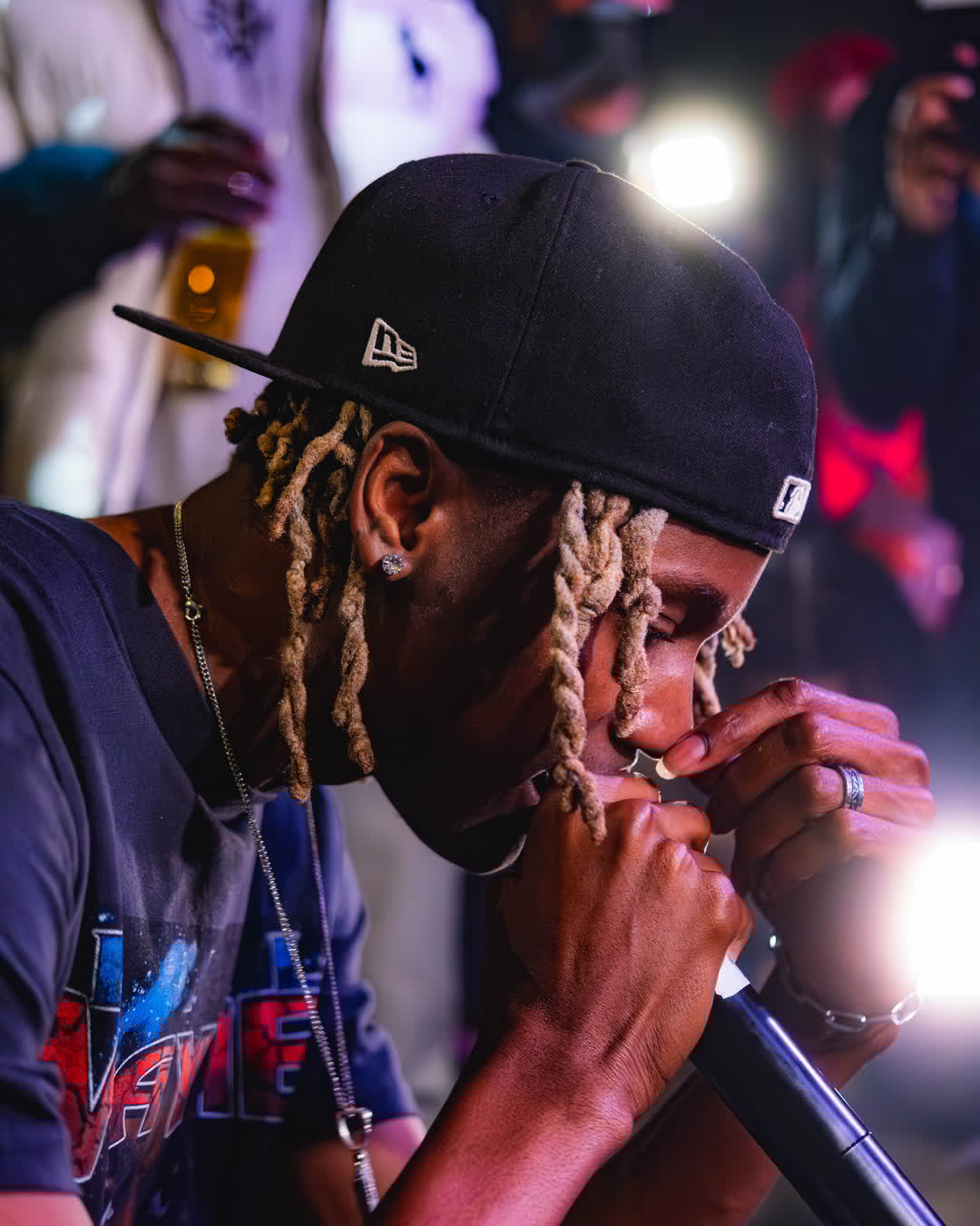 A musical artist with dreadlocks and a black fitted cap holding a microphone close to his face during a live, intensely lit performance.