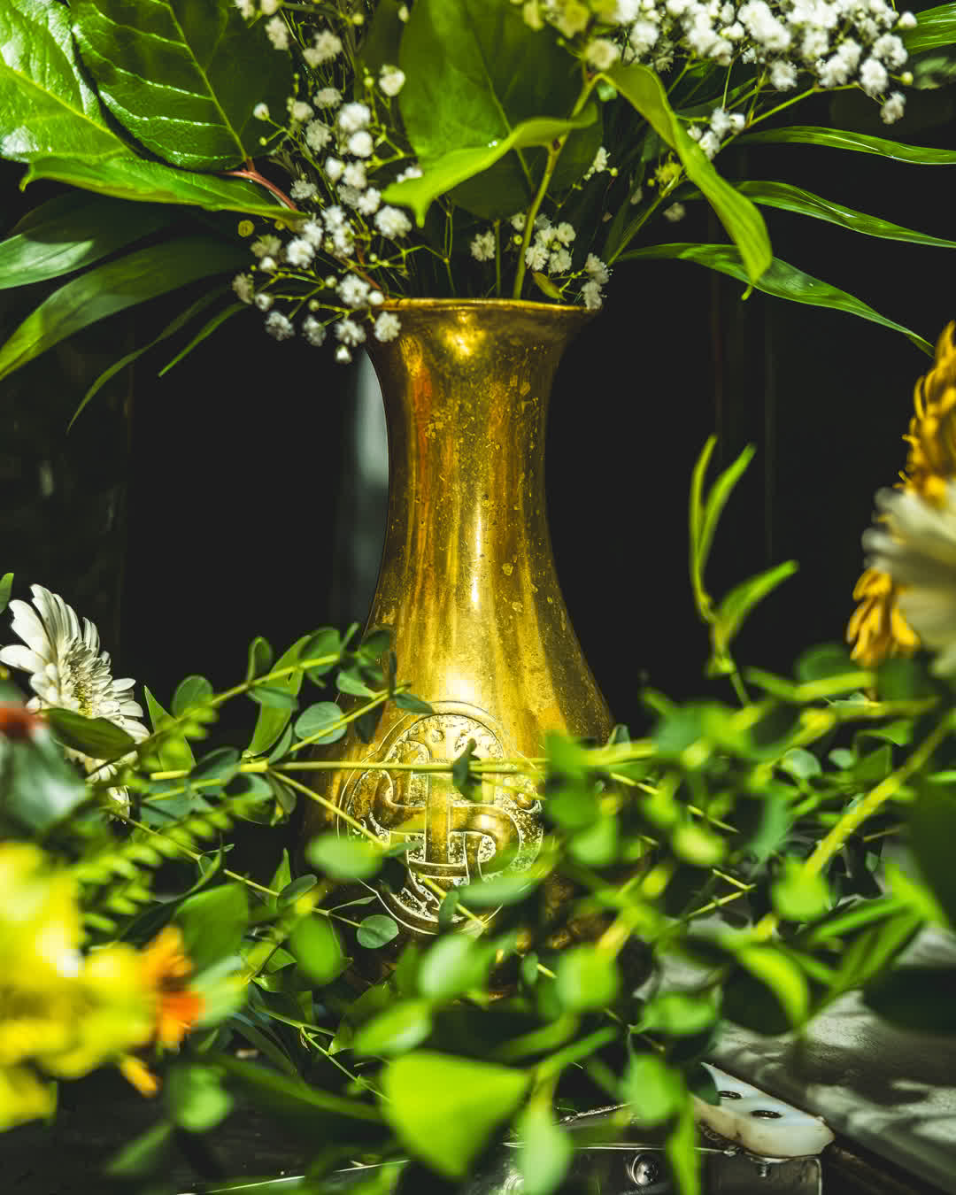 A golden, ornate vase holding white baby's breath and green foliage, partially obscured by vibrant green foreground leaves.